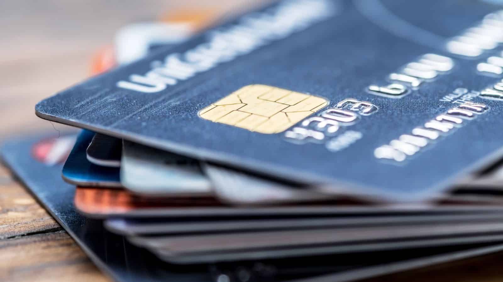 A close-up of a stack of credit cards with visible chip technology, showing the embossed numbers and details on the top card, resting on a wooden surface.