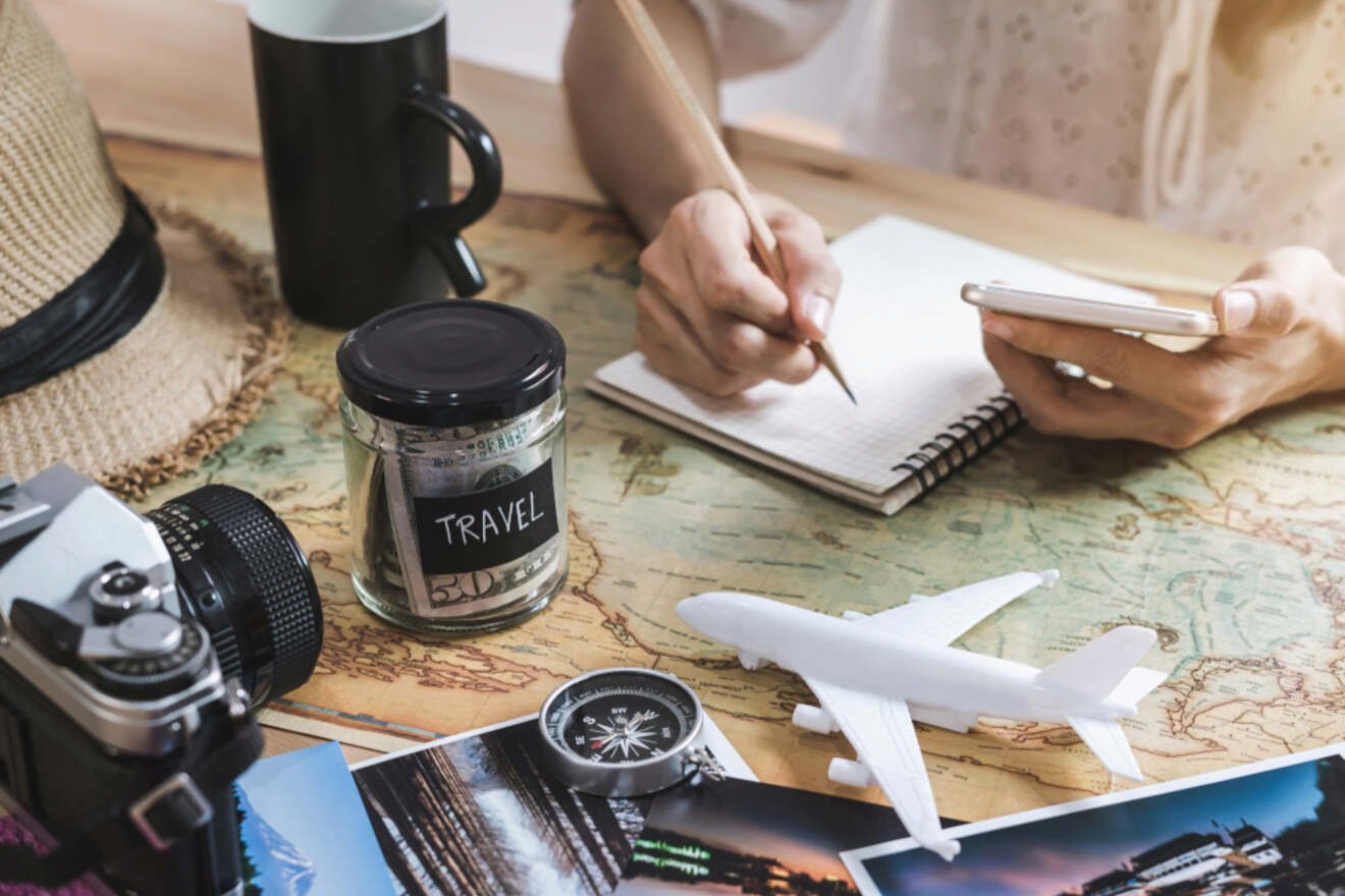 A person is planning a trip, writing in a notebook with travel items such as a map, camera, toy airplane, compass, and jar labeled "Travel" on the table.