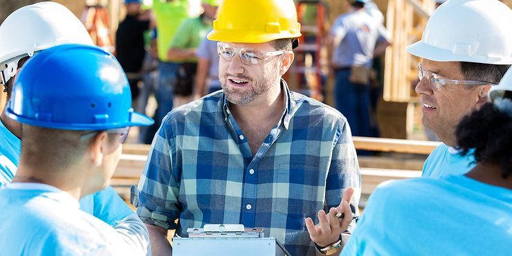 A construction foreman in a yellow hard hat leads a team briefing on a sunny job site.