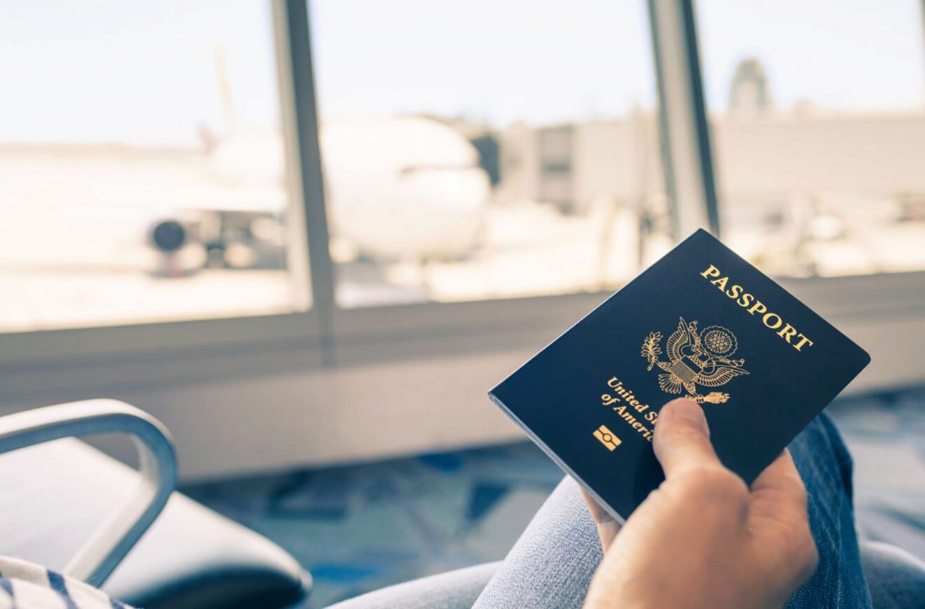 Close-up of a person holding a United States passport with an airplane visible through the airport window in the background