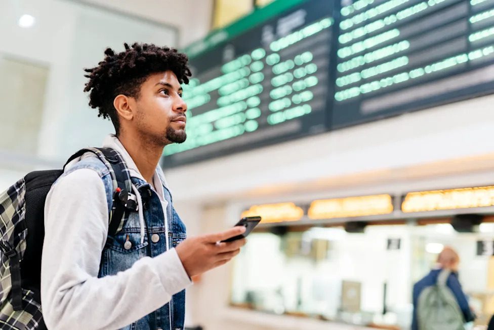 A man holding a mobile phone at the airport, looking at flight boards