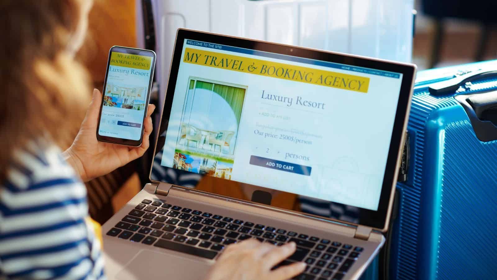 A person looks at a travel booking website for a luxury resort on a laptop and smartphone. A blue suitcase and another silver suitcase are nearby, suggesting a travel or vacation setting.