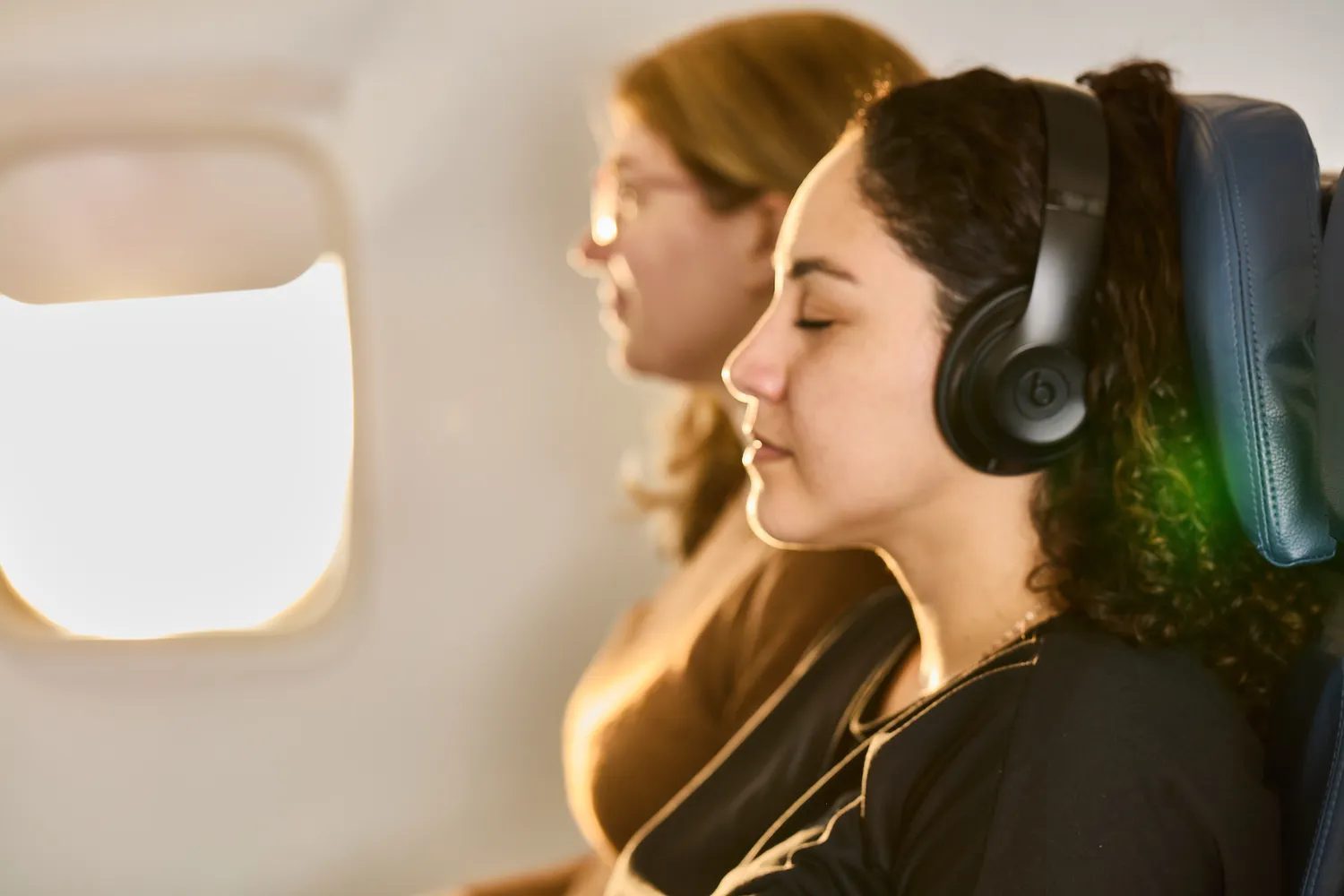 Two passengers seated on an airplane one wearing headphones and the other appearing relaxed near a window