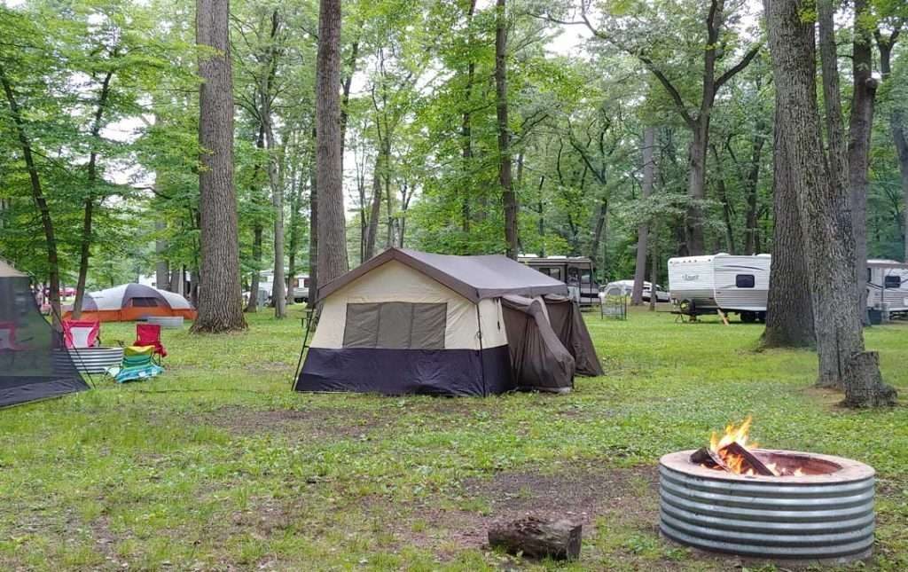 Campsite at Bay City State Park featuring a large beige and brown tent, camping chairs, a campfire, and RVs in the background.