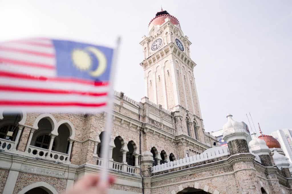Person holding a Malaysian flag in front of a historic building with a clock tower